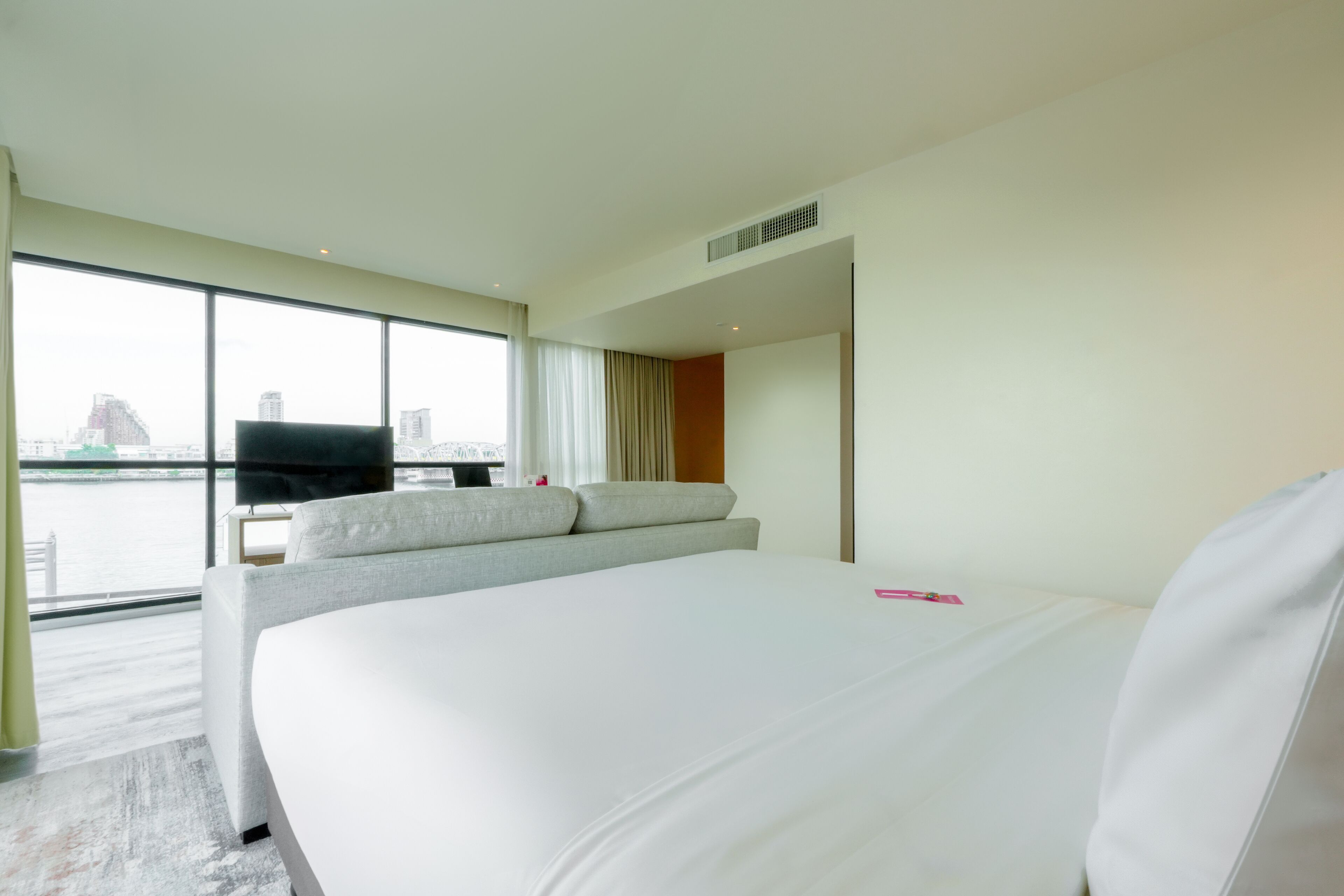 Ein Hotelzimmer mit einem großen Bett, einem Fernseher und Blick auf eine Stadtlandschaft durch große Fenster.