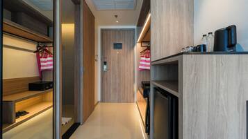 A modern hotel room with a wooden wardrobe, a built-in shelf, and a small kitchen area with a coffee machine.