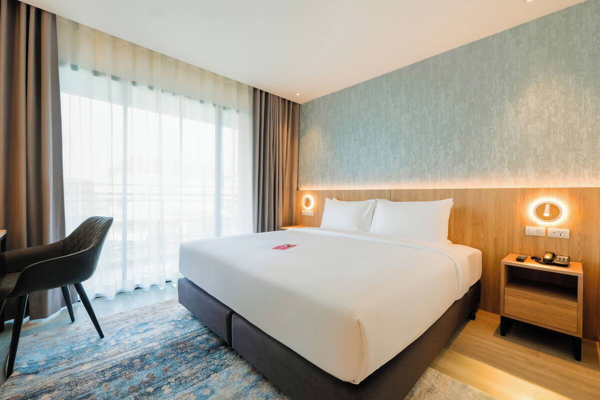 A modern hotel room with a large bed, a desk, and a chair. There is a nightstand with a lamp and a small table.