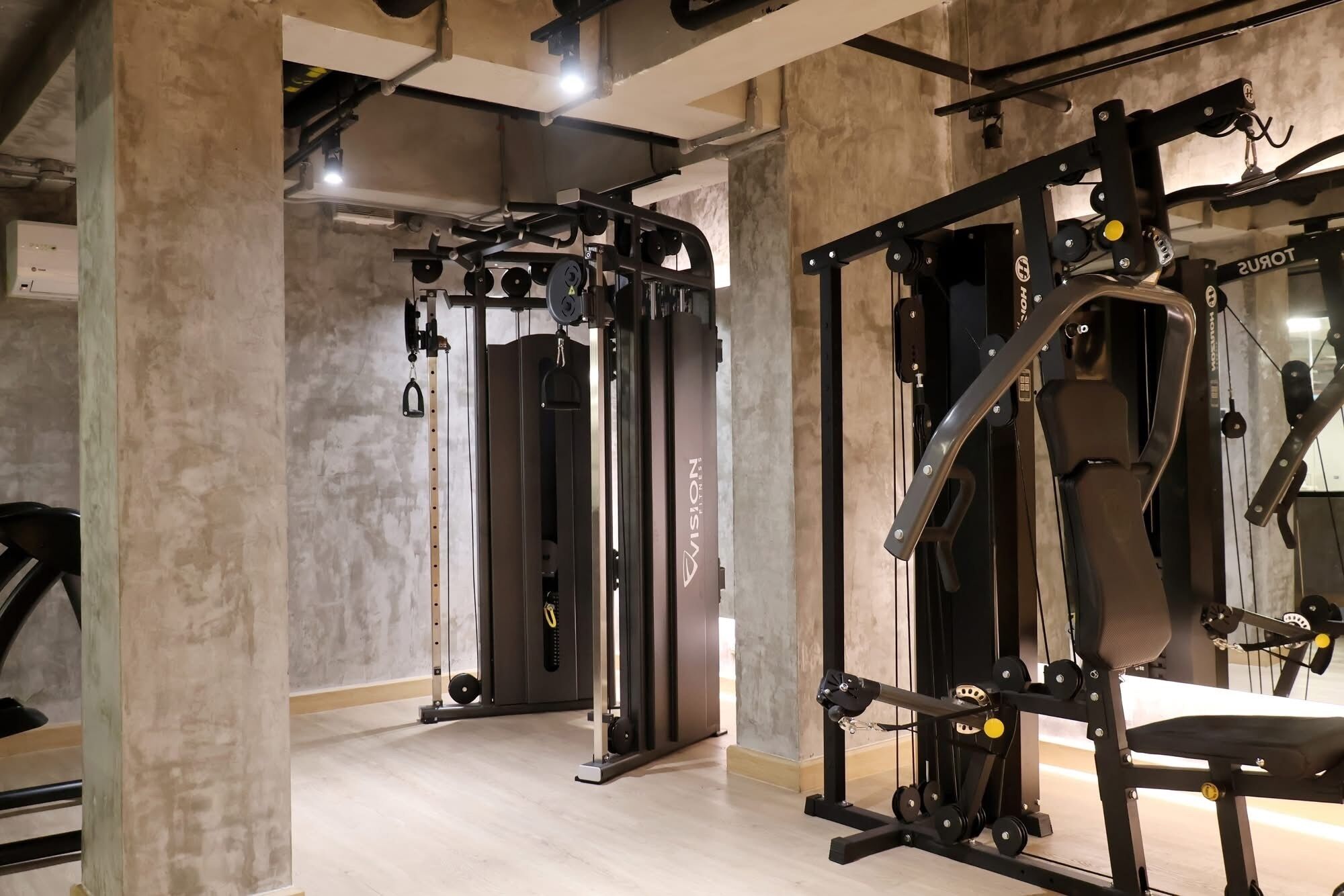 Un gimnasio con equipo de levantamiento de pesas, pilares de hormigón y suelo de madera.