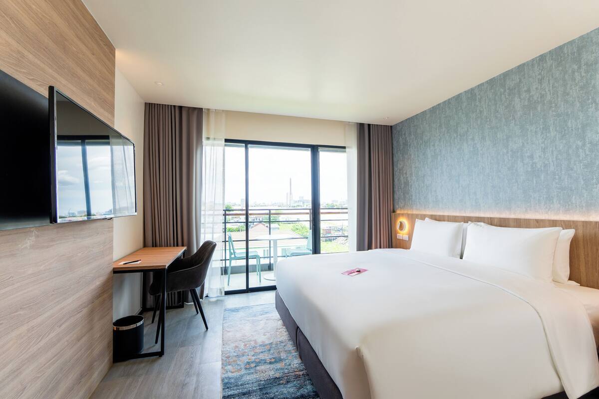 A modern hotel room with a large bed, a desk with a chair, a flat-screen TV mounted on the wall, and a balcony with a view of the city.