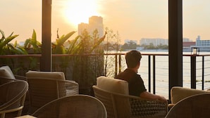 A person seated on wicker chairs overlooking a body of water with a cityscape in the background. - TARA Bangkok Riverside (Formerly GLOW Bangkok Riverside) (Bangkok)