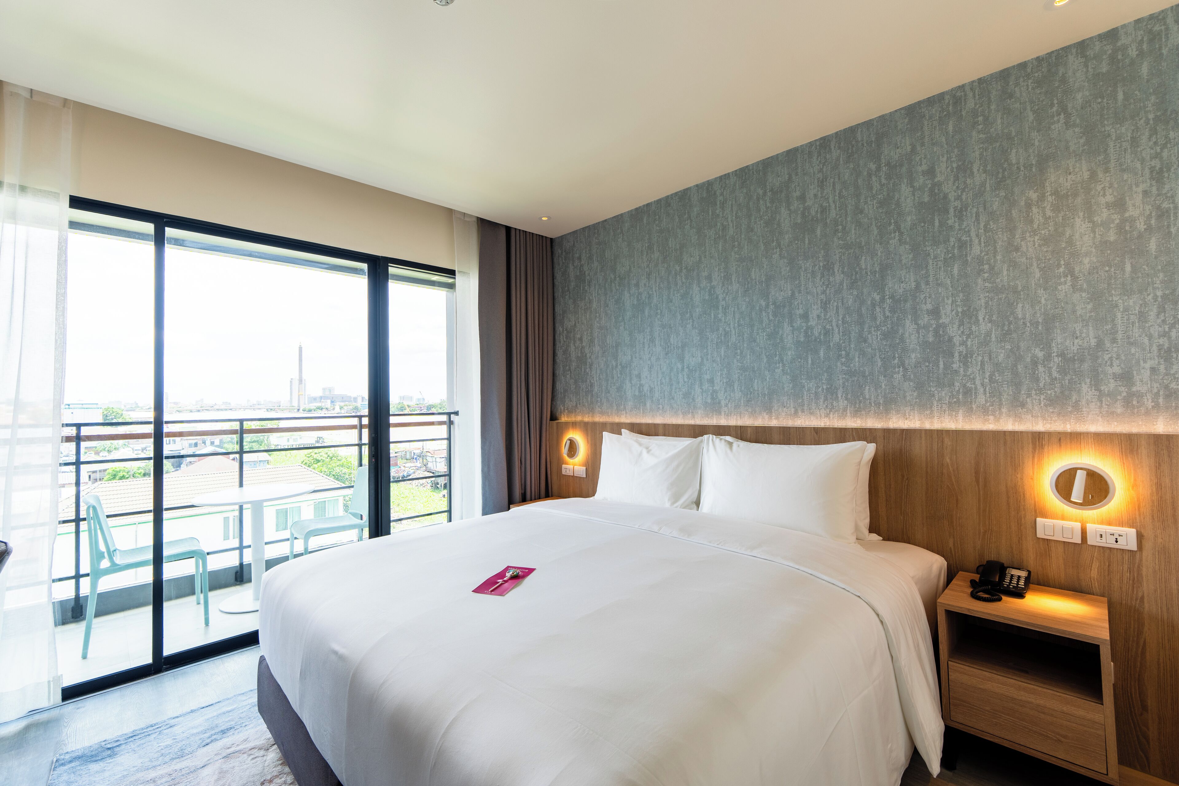Ein modernes Hotelzimmer mit einem großen Bett, einem Nachttisch mit Telefon, einem kleinen Schreibtisch und Blick auf die Stadt durch große Fenster.