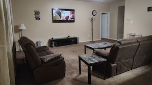 Una sala de estar con un televisor de pantalla plana grande, un ventilador de techo, un perchero y una puerta al fondo.