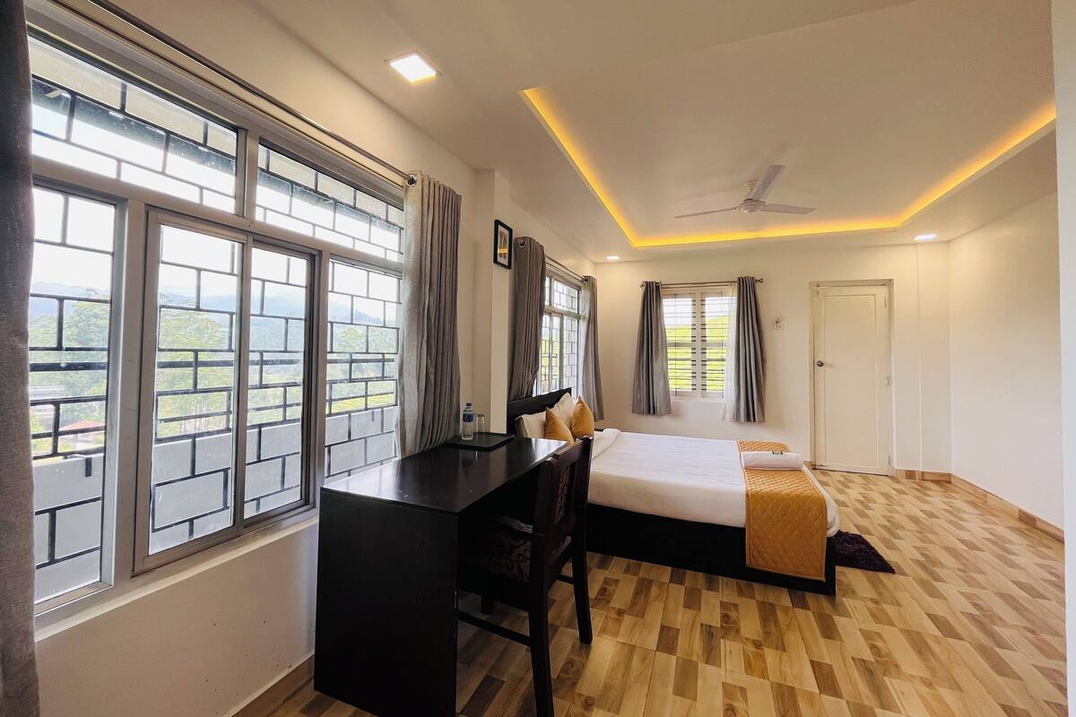 A hotel room with a bed, desk, and large windows offering a view of the outdoors.