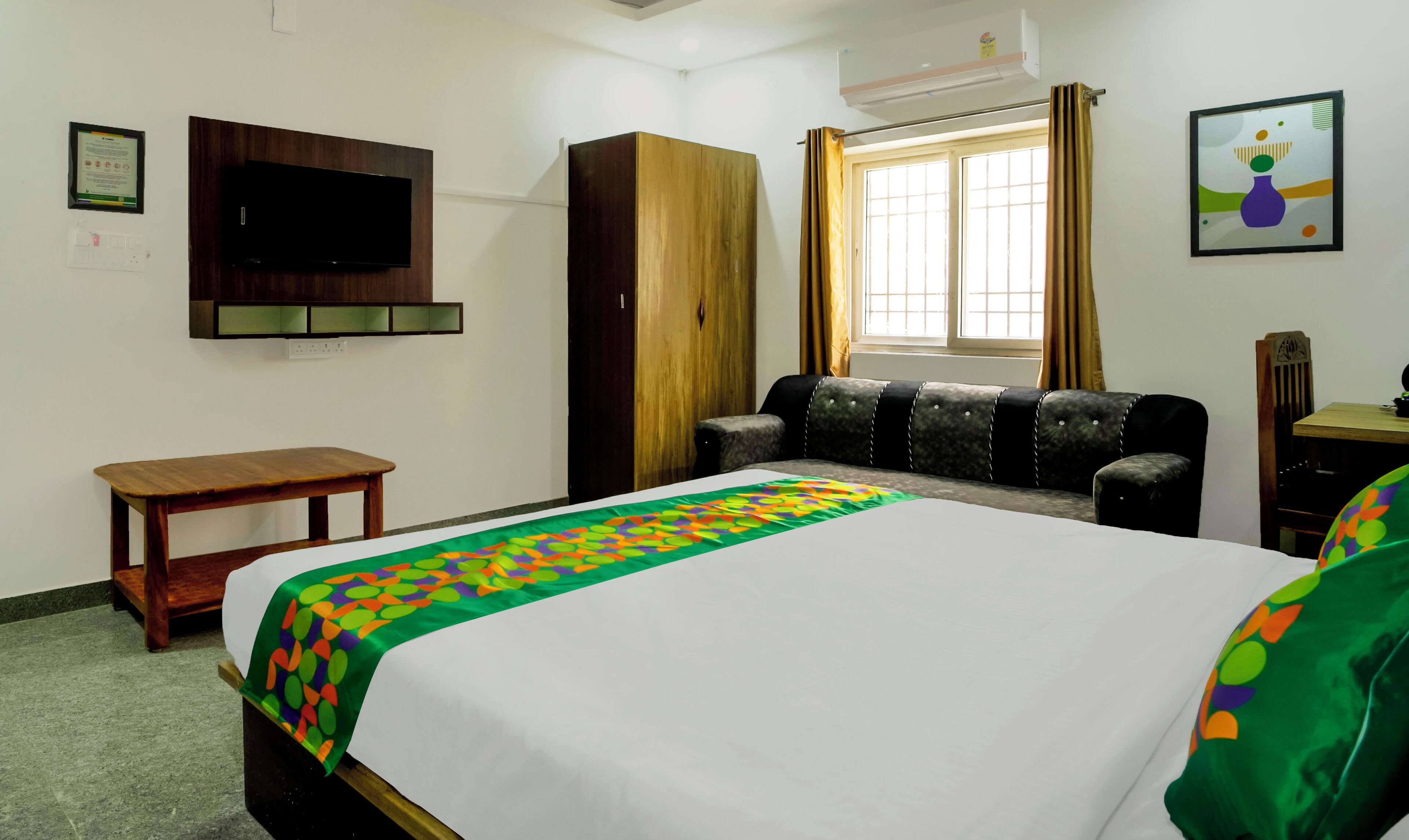 Une chambre d’hôtel avec un lit, un téléviseur fixé au mur, une armoire en bois, une fenêtre avec des rideaux, une petite table et une chaise.