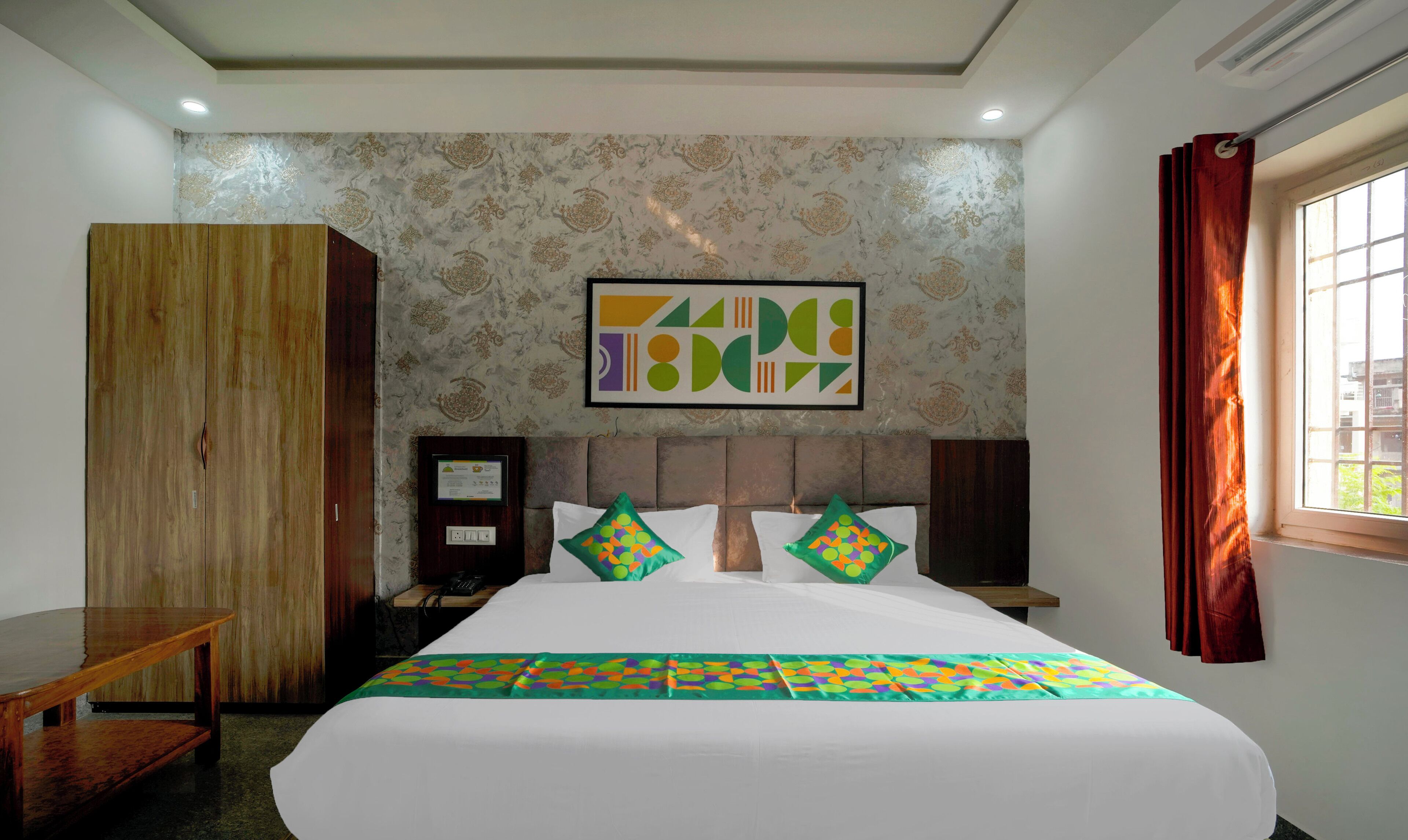 A hotel room with a bed, a wooden wardrobe, a desk, a telephone, and a window with curtains.
