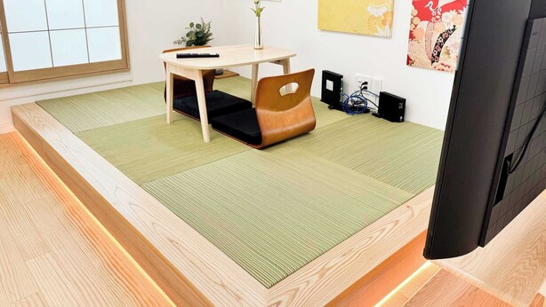 A room with a tatami mat floor, a low wooden bed frame, a small table with a chair, a TV, and a wall-mounted speaker.