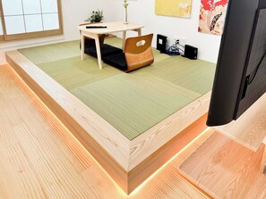 A room with a tatami mat floor, a low wooden bed frame, a small table with a chair, a TV, and a wall-mounted speaker. - Modern Takadanobaba House near Cherry Blossoms & Stations (Shinjuku City)
