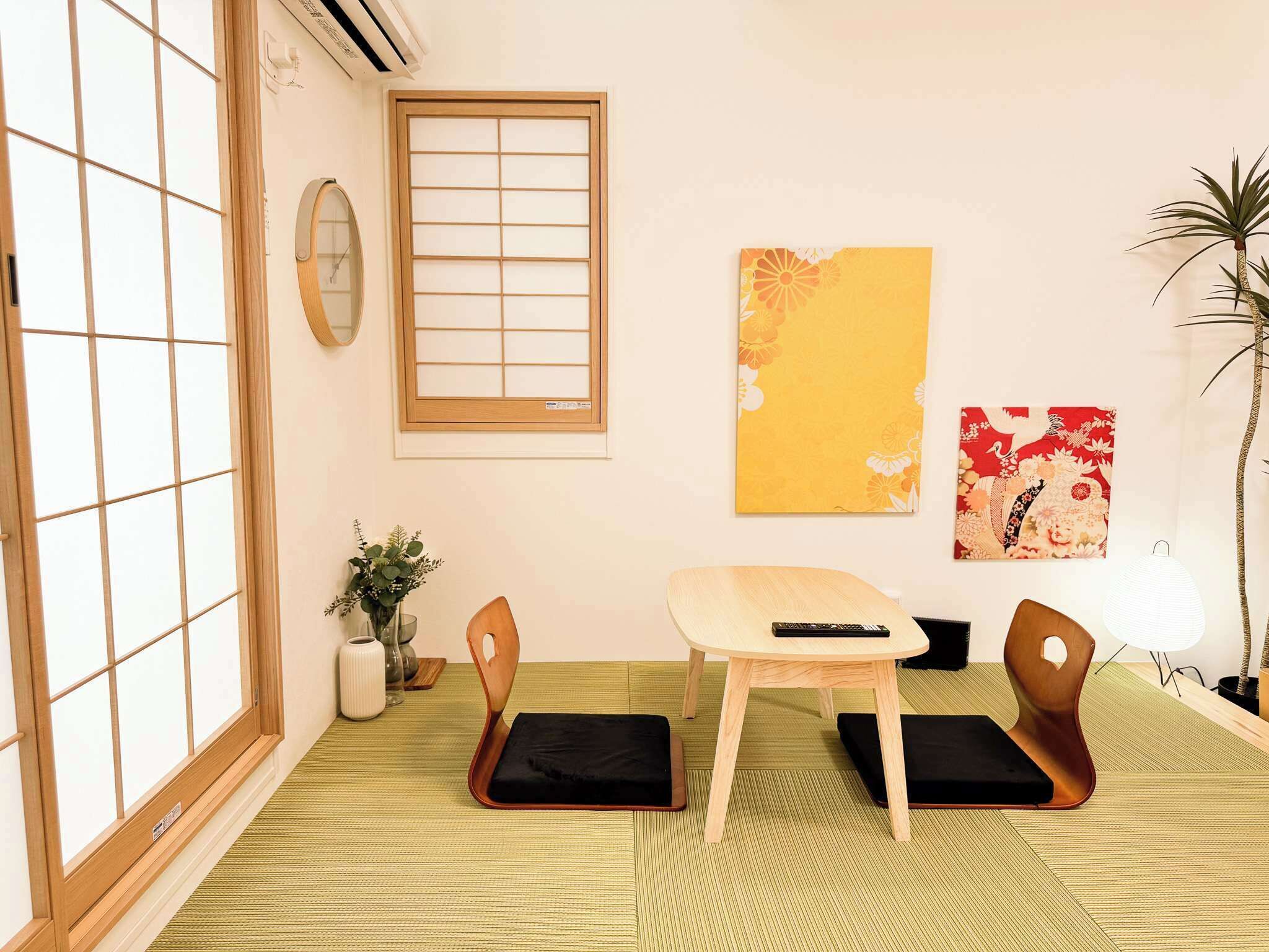 A traditional Japanese room with tatami flooring, a low table, two wooden chairs, a small potted plant, and artwork on the walls.