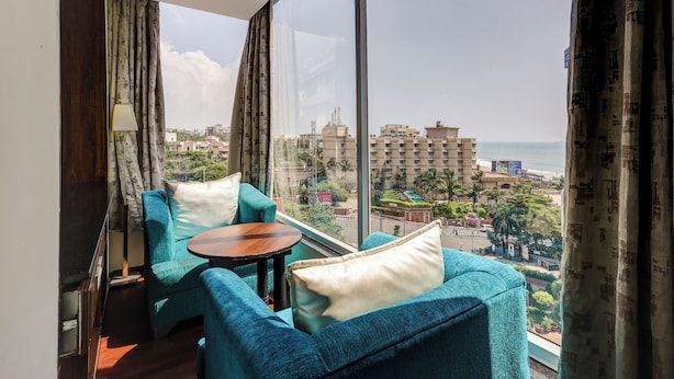 Een balkon met twee blauwe fauteuils, een klein rond tafeltje en uitzicht op een stadsbeeld en strand.