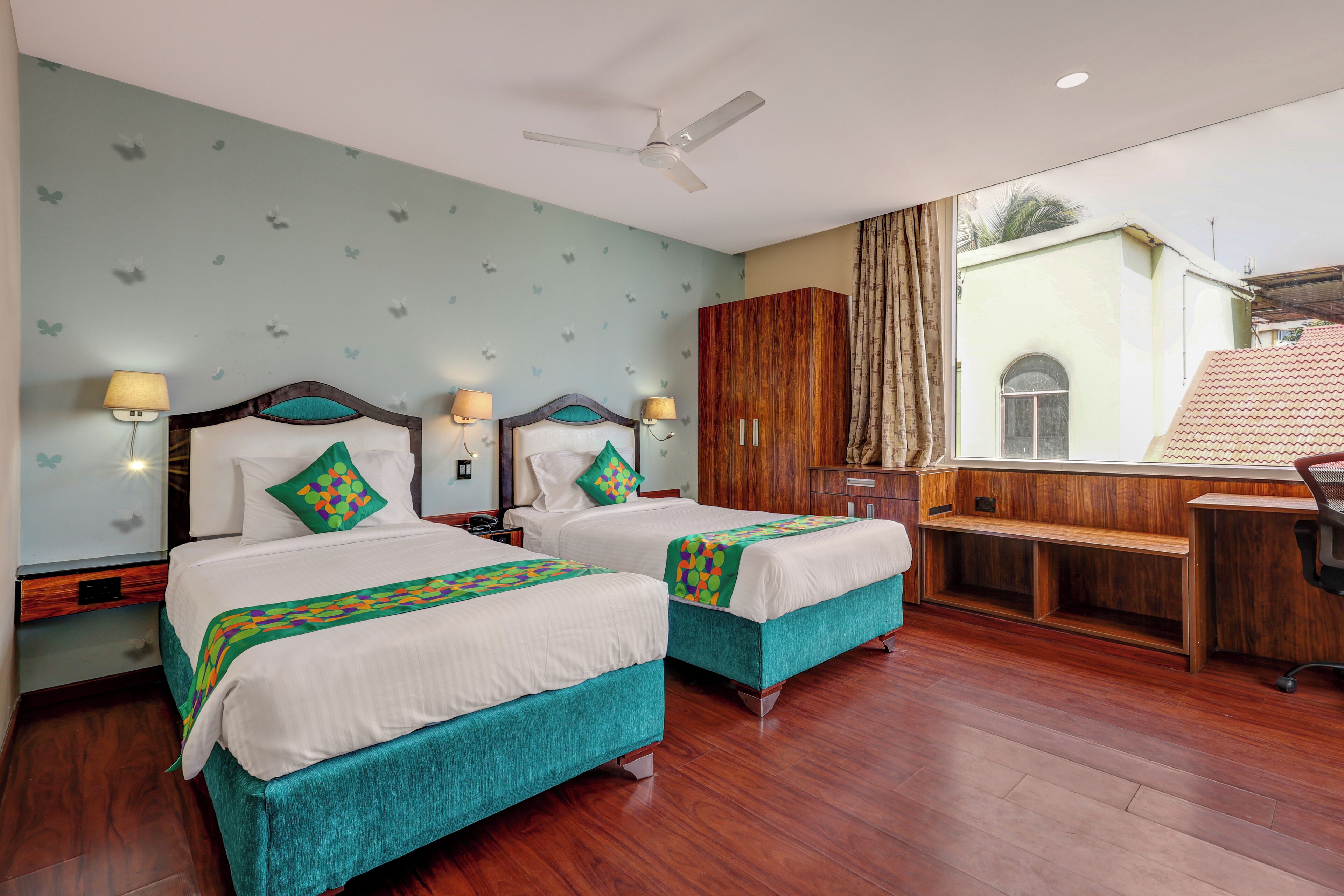 A hotel room with two beds, a desk, a chair, a ceiling fan, and a window with curtains.