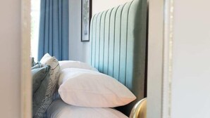 Une chambre à coucher avec un lit, des oreillers, une table de chevet avec une plante et une fenêtre avec des rideaux.
