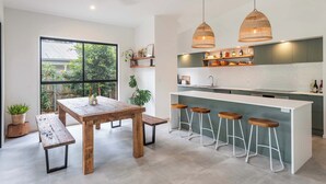 A modern kitchen with a wooden table, bar stools, pendant lights, and a view through a large window.