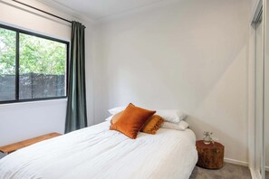 Een slaapkamer met een bed, een raam met gordijnen, een nachtkastje en een klein houten krukje.