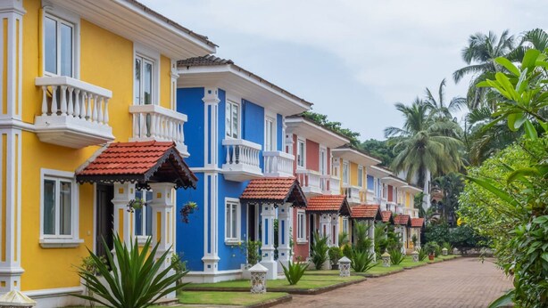 A row of colorful colonial-style buildings with balconies and red-tiled roofs, lined with lush greenery and palm trees. - 4BR Villa With Pool 10 Mins From Calangute Beach (Calangute)