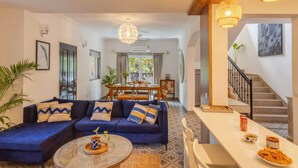 A modern living room with a blue sofa set, a dining table, and a staircase leading to an upper floor. - 4BR Villa With Pool 10 Mins From Calangute Beach (Calangute)