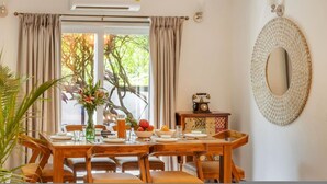 A dining area with a wooden table set for four, a vase with flowers, and a fruit bowl. - 4BR Villa With Pool 10 Mins From Calangute Beach (Calangute)
