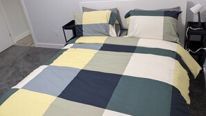 A bed with a checkered bedspread in neutral tones, a bedside table with a lamp, and a plain white wall. - President's Choice: Spacious 1-Bedroom Basement Suite with Fireplace (Edmonton)