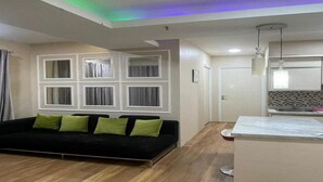 A modern living room with a black sofa, green cushions, a wooden floor, and a kitchen area with a white countertop and a hanging light fixture. - Hotel O Grand Asia Afrika Apartment By Qr Property (Bandung)