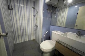 A bathroom with a striped tile wall, a white toilet, a sink with a mirror, and a shower with a handheld showerhead. - Hotel O Grand Asia Afrika Apartment By Qr Property (Bandung)
