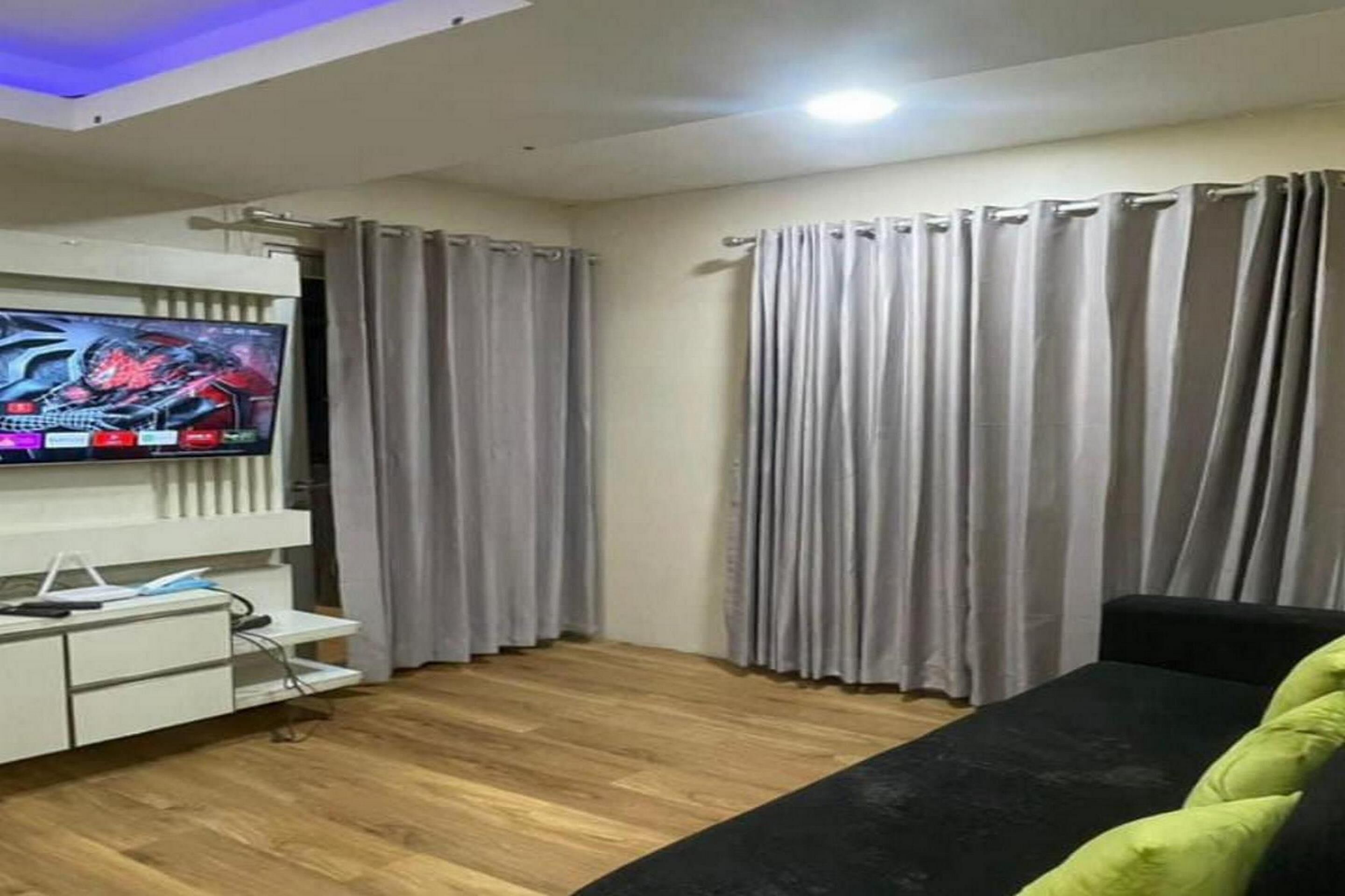 A modern living room with a flat-screen TV, a white entertainment unit, a black sofa, and grey curtains.