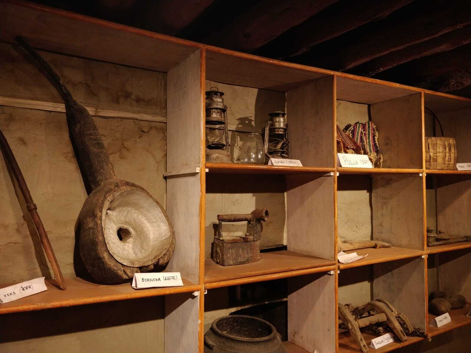 A display of historical artifacts in a museum setting, including wooden shelves with labeled items such as a large wooden wheel, a lantern, and various tools.