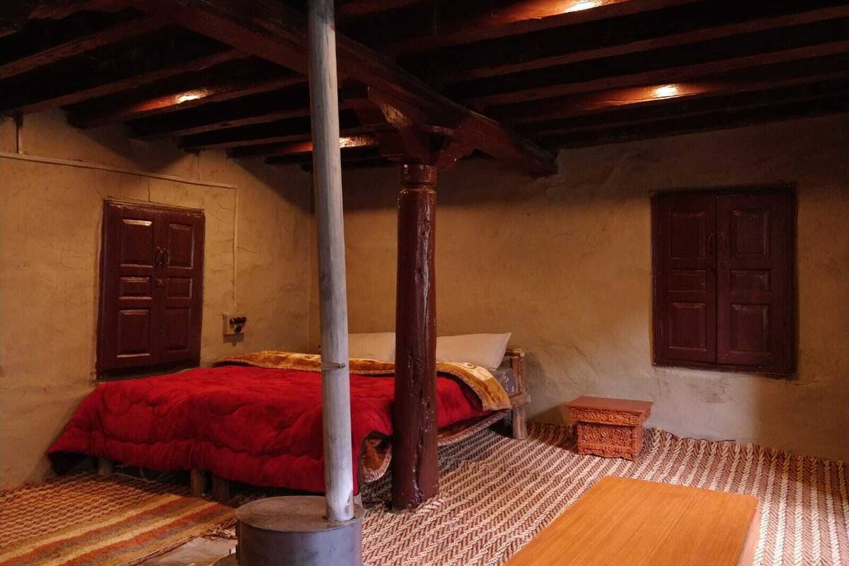A room with a bed, a wooden pole, a small table, and a woven rug.