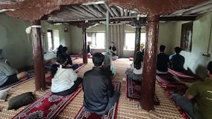 Une pièce où un groupe de personnes est assis sur des tapis à motifs, entouré de piliers en bois et coiffée d’un plafond simple et sans ornements.