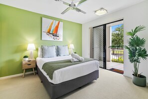 Ein Schlafzimmer mit einem Bett, zwei Nachttischen mit Lampen, einem Deckenventilator und einem großen Fenster mit Blick auf Grünflächen.