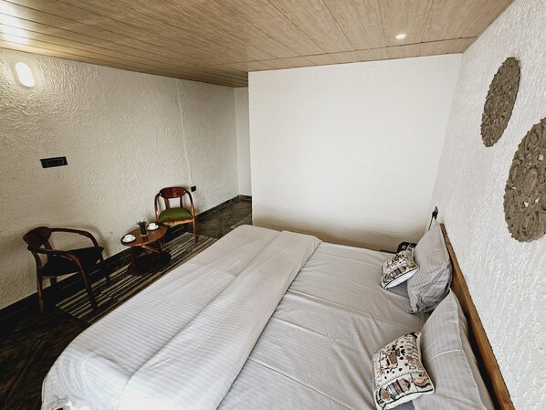 A bedroom with a large bed, a wooden chair, a small table with a cup and saucer, and decorative wall art.
