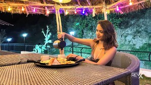 A person pouring a drink into a cup at a table with a wicker chair, a tray with food and drinks, and a thatched roof overhead. - Anandvan Jungle Resort (Rishikesh)