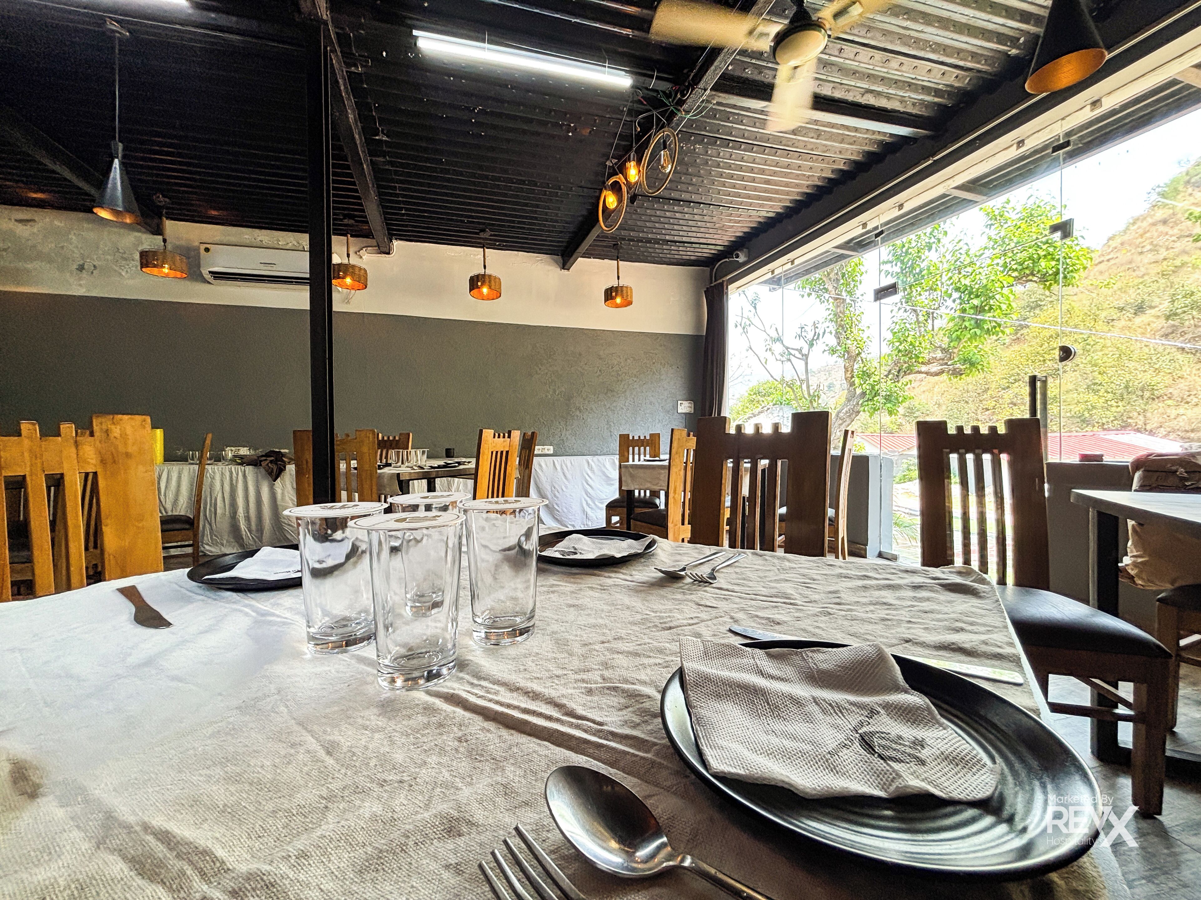 Un área de comedor al aire libre con mesas dispuestas para dos, con platos de metal, cubiertos y vasos transparentes.