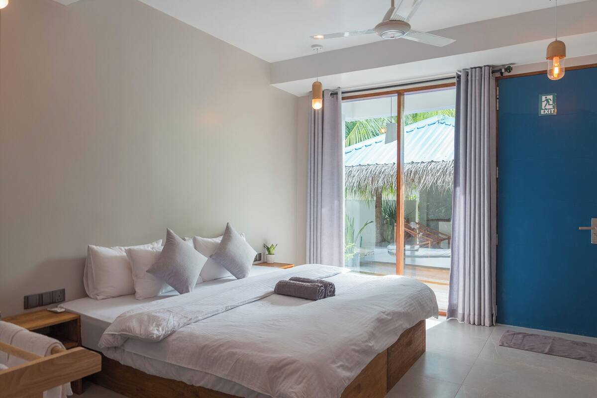 A bedroom with a large bed, a nightstand, a ceiling fan, and a view of the outdoors through a glass door.