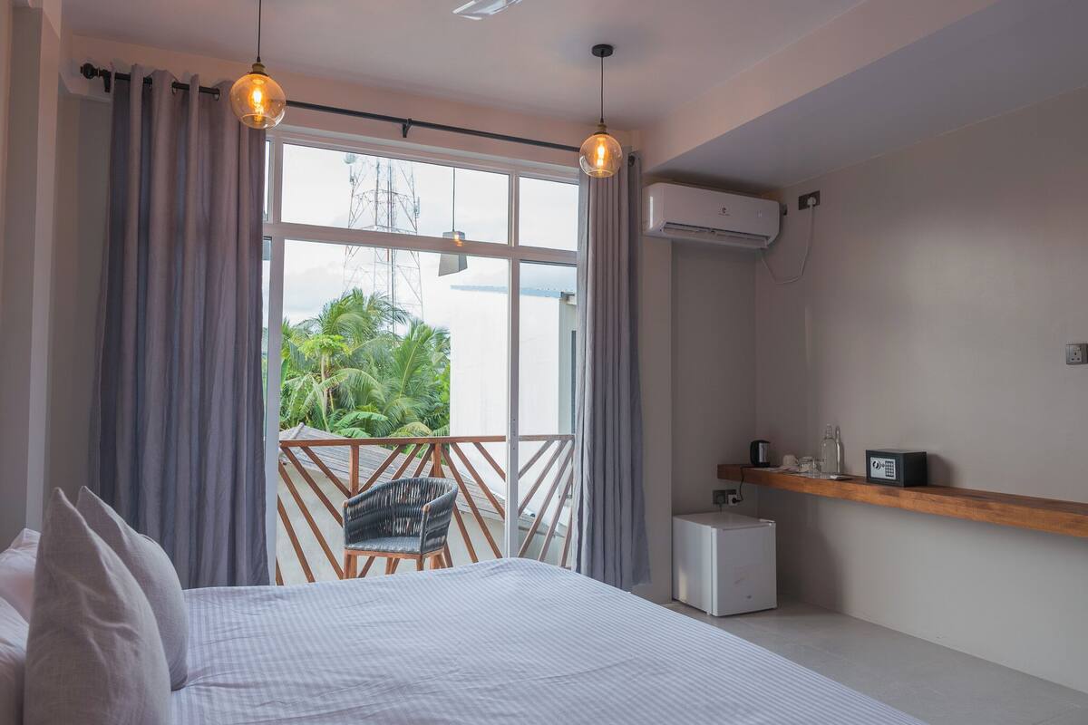 A bedroom with a bed, a chair, a small table, an air conditioning unit, and a view of greenery outside.