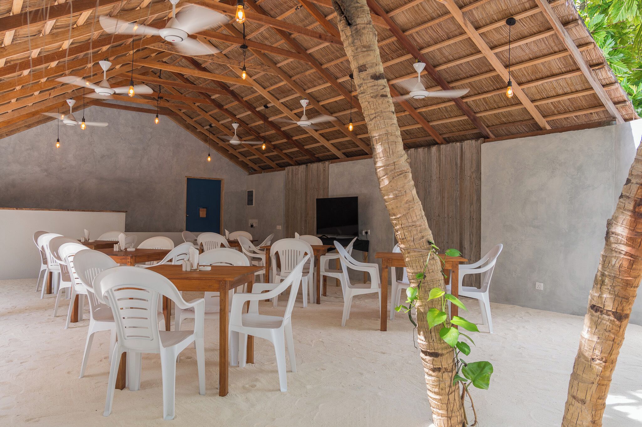 A covered outdoor dining area with white plastic chairs and tables, ceiling fans, and a TV screen on the wall.