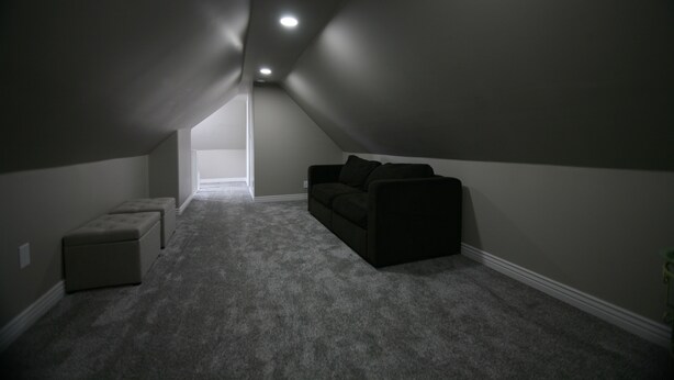 Un ático tenuemente iluminado con un sofá y reposapiés de color oscuro, piso alfombrado y una puerta visible que da a otra habitación.