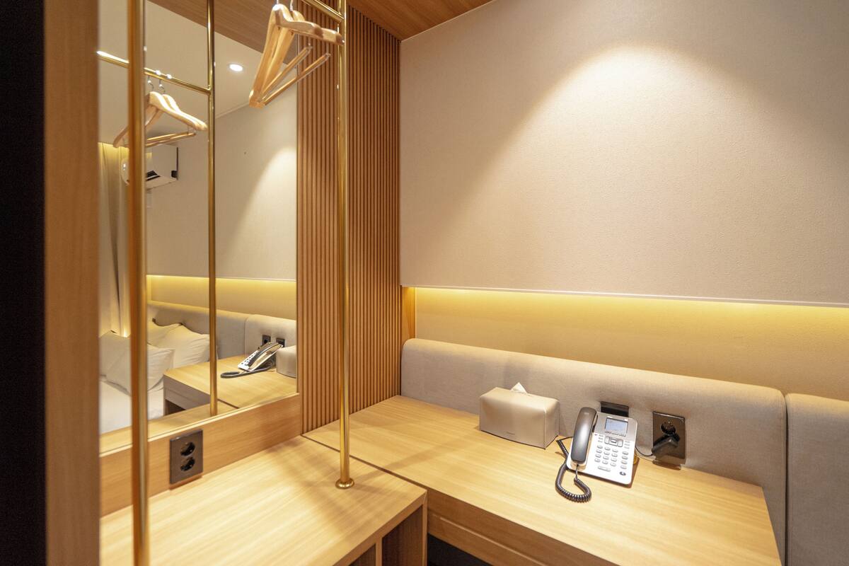 Une chambre d’hôtel avec une coiffeuse en bois, un miroir, une table de chevet avec un téléphone et un banc rembourré.