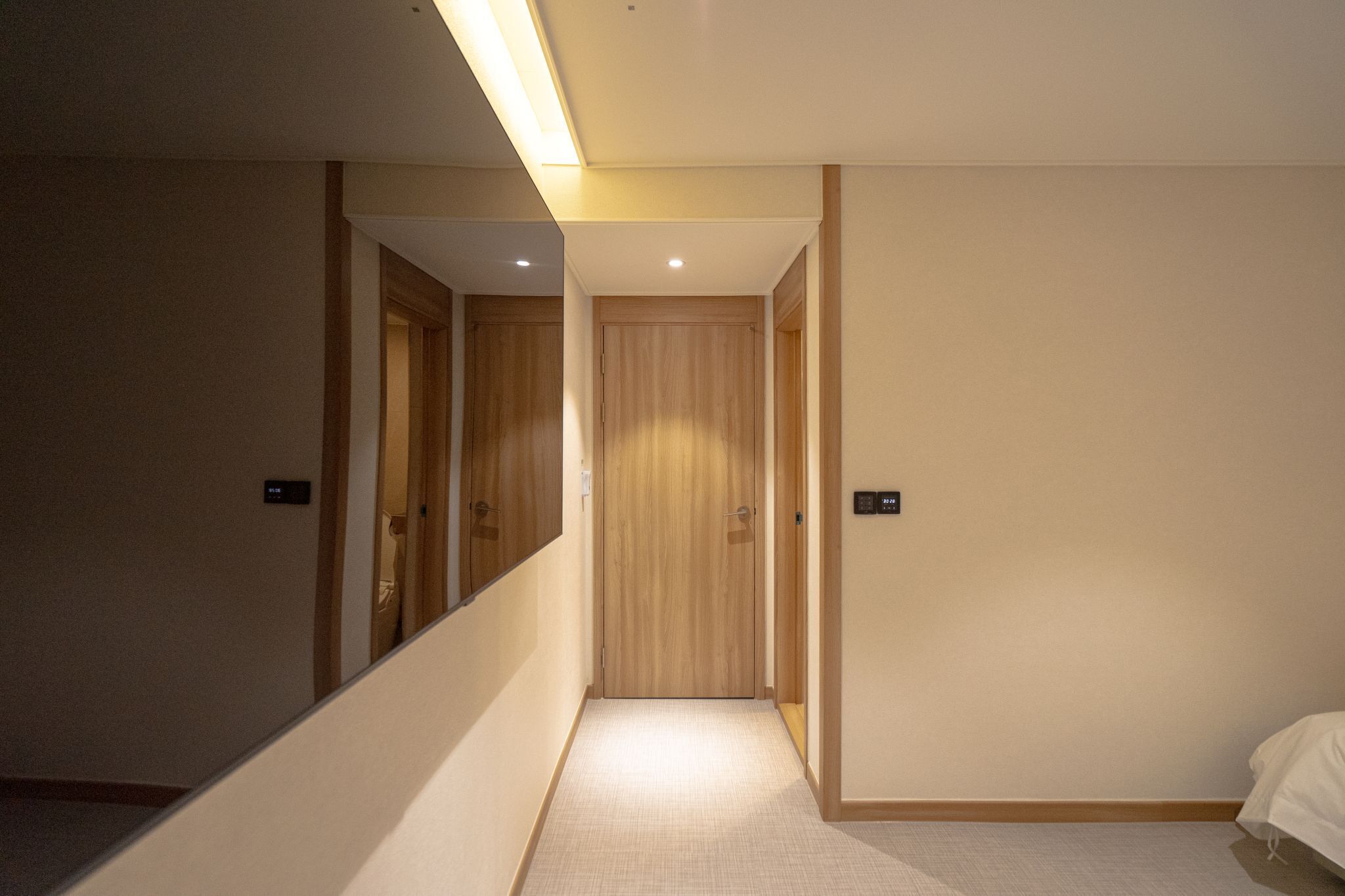 A hotel hallway with a wooden door, a mirror, and a bed in the adjacent room.