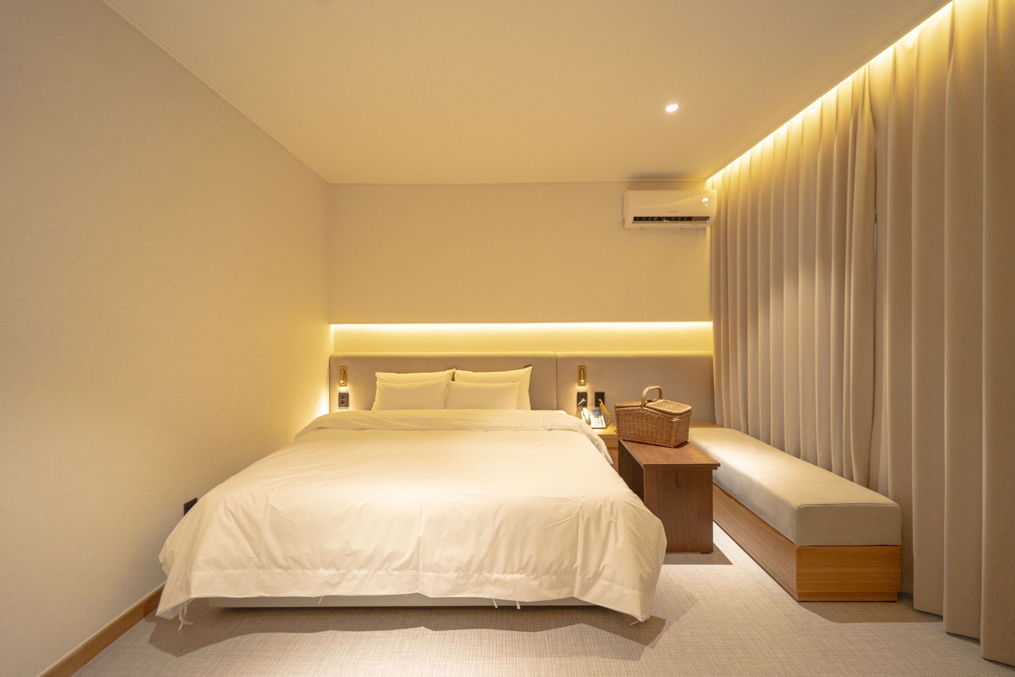 A modern hotel room with a large bed, bedside tables, a bench, and a wall-mounted air conditioning unit.