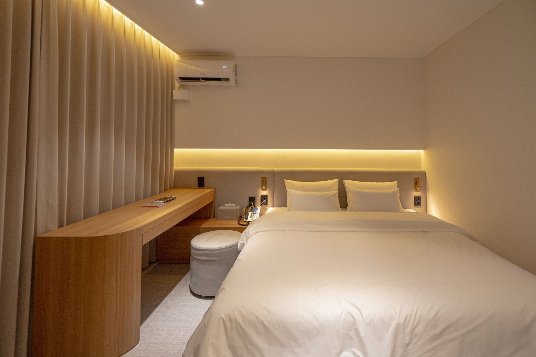 A hotel room with a large bed, a wooden desk, a trash bin, and a wall-mounted air conditioner.