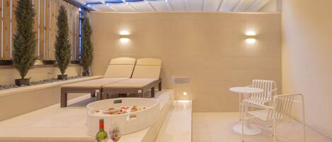 Un espace spa moderne comprenant une table de massage, un coin salon avec une petite table et des chaises, ainsi qu’un arrangement floral décoratif.