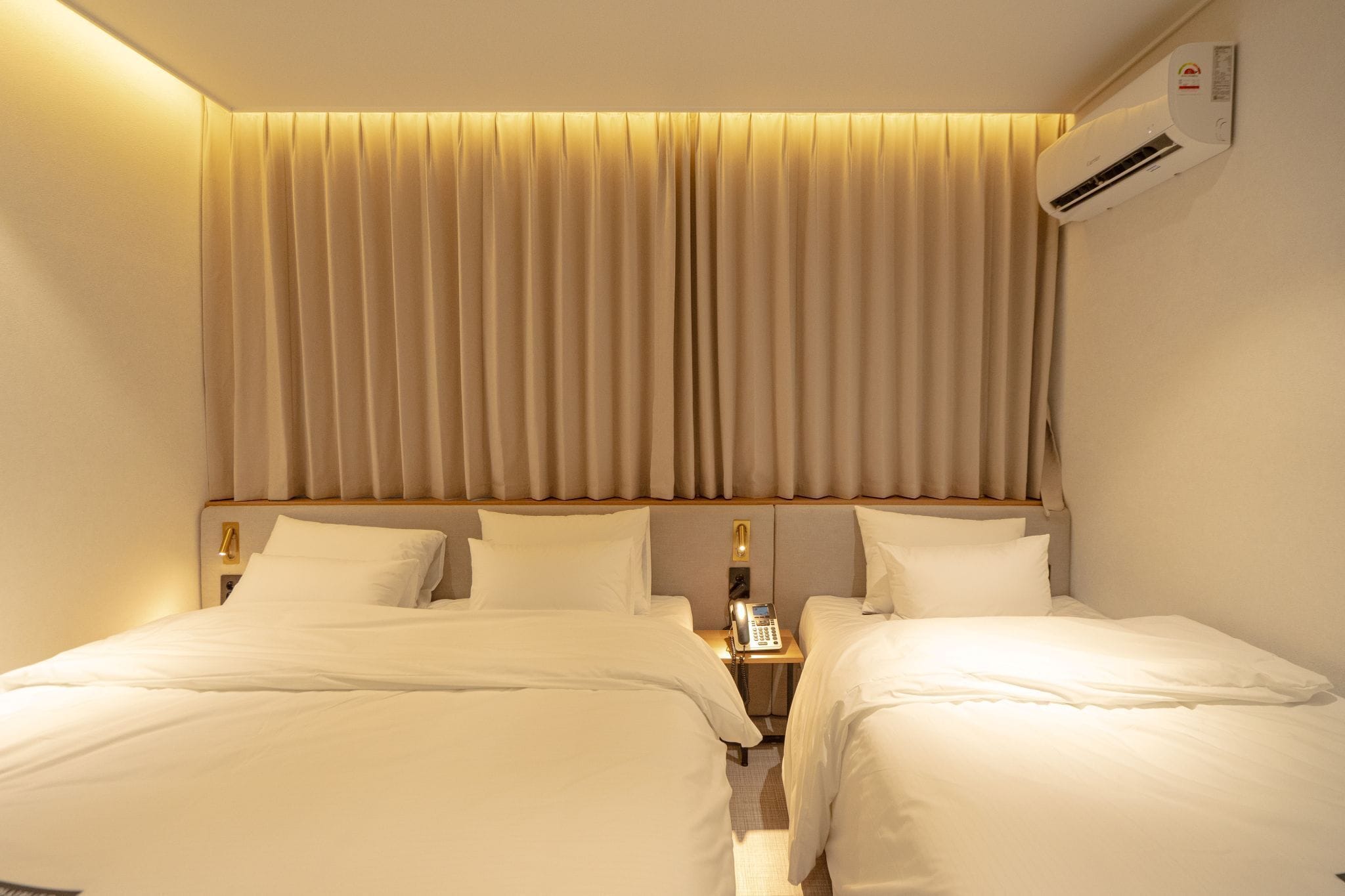 Una habitación de hotel con dos camas, una mesita de noche con teléfono y un equipo de aire acondicionado en la pared.