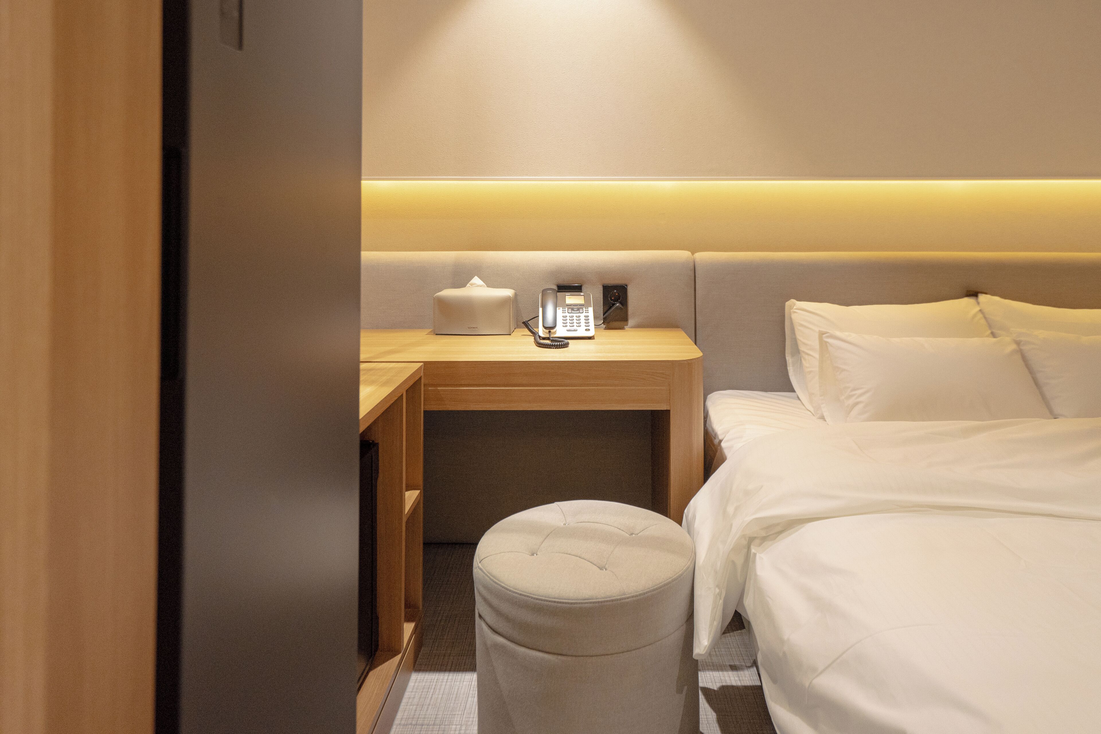 A hotel room with a bed, a bedside table with a lamp, a telephone, and a small ottoman.