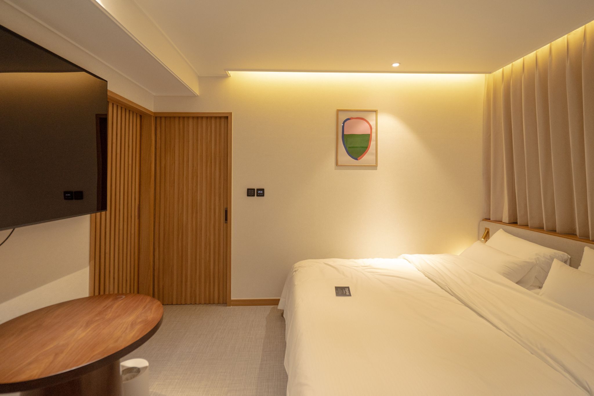 A hotel room with a bed, a wooden desk, a flat-screen TV mounted on the wall, and a painting on the wall.