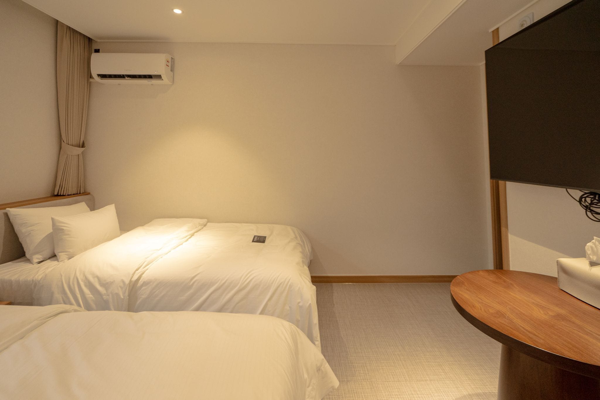 Une chambre d’hôtel avec deux lits, une table de chevet en bois, un climatiseur fixé au mur et un téléviseur à écran plat.