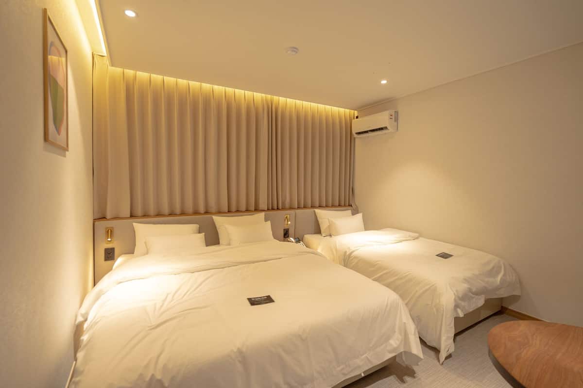 Une chambre d’hôtel avec deux lits, une tête de lit en bois, un climatiseur fixé au mur et un petit banc en bois.