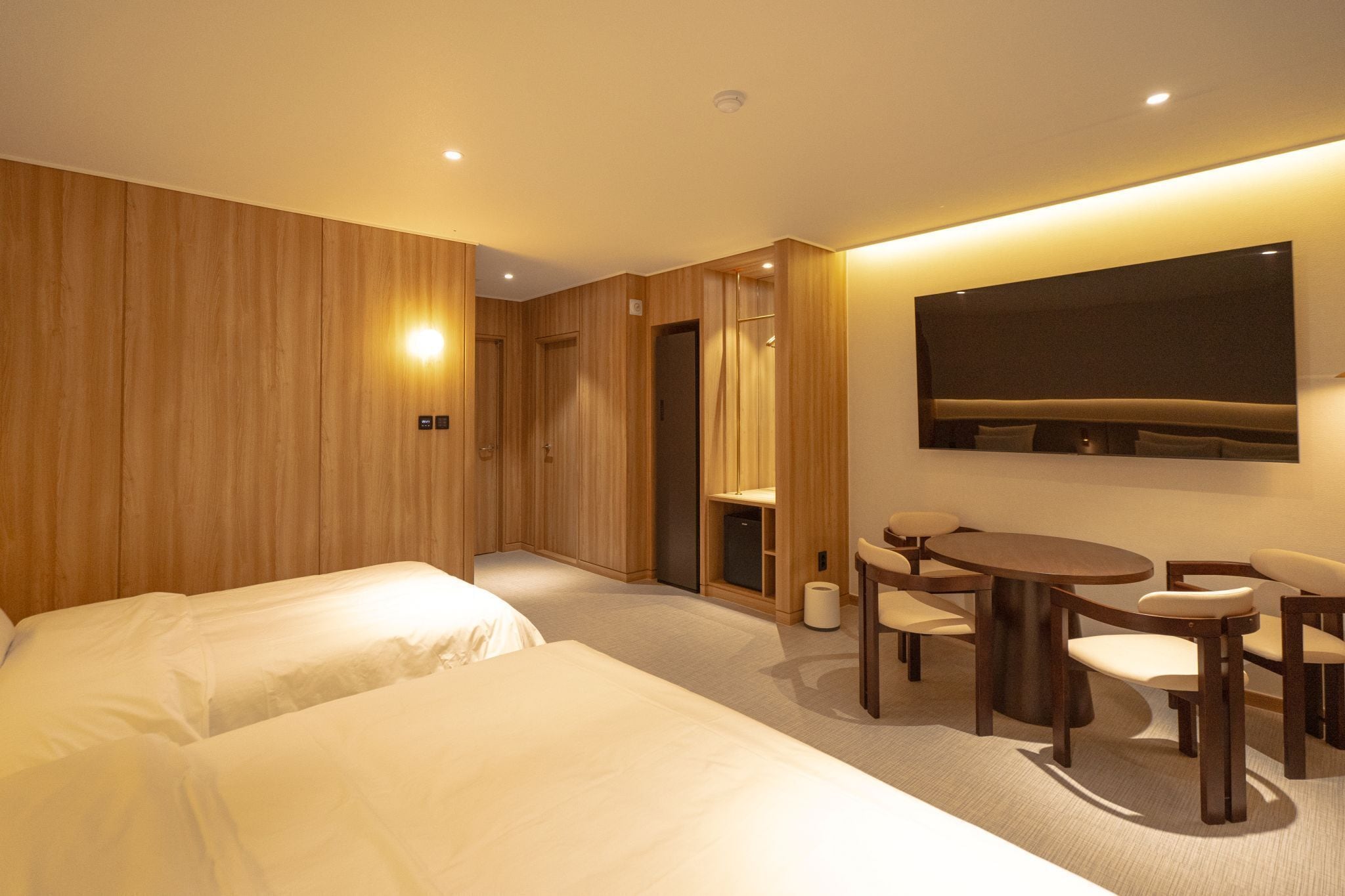 Habitación de hotel con dos camas, una mesa de comedor con sillas, un televisor de pantalla plana en la pared y un panel de pared de madera.