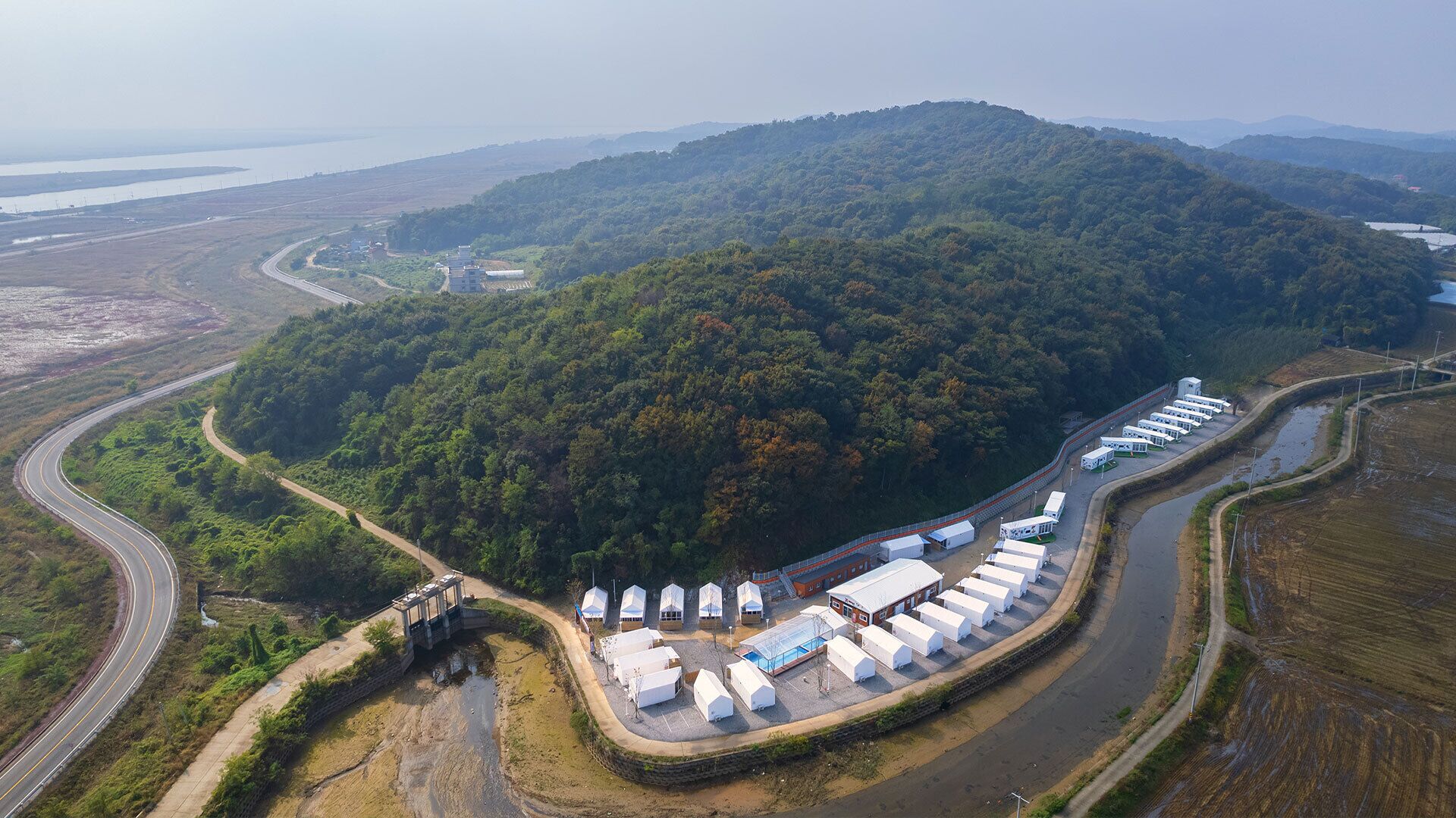 An aerial view of a campsite with tents arranged in rows, surrounded by a forested area and a winding road.