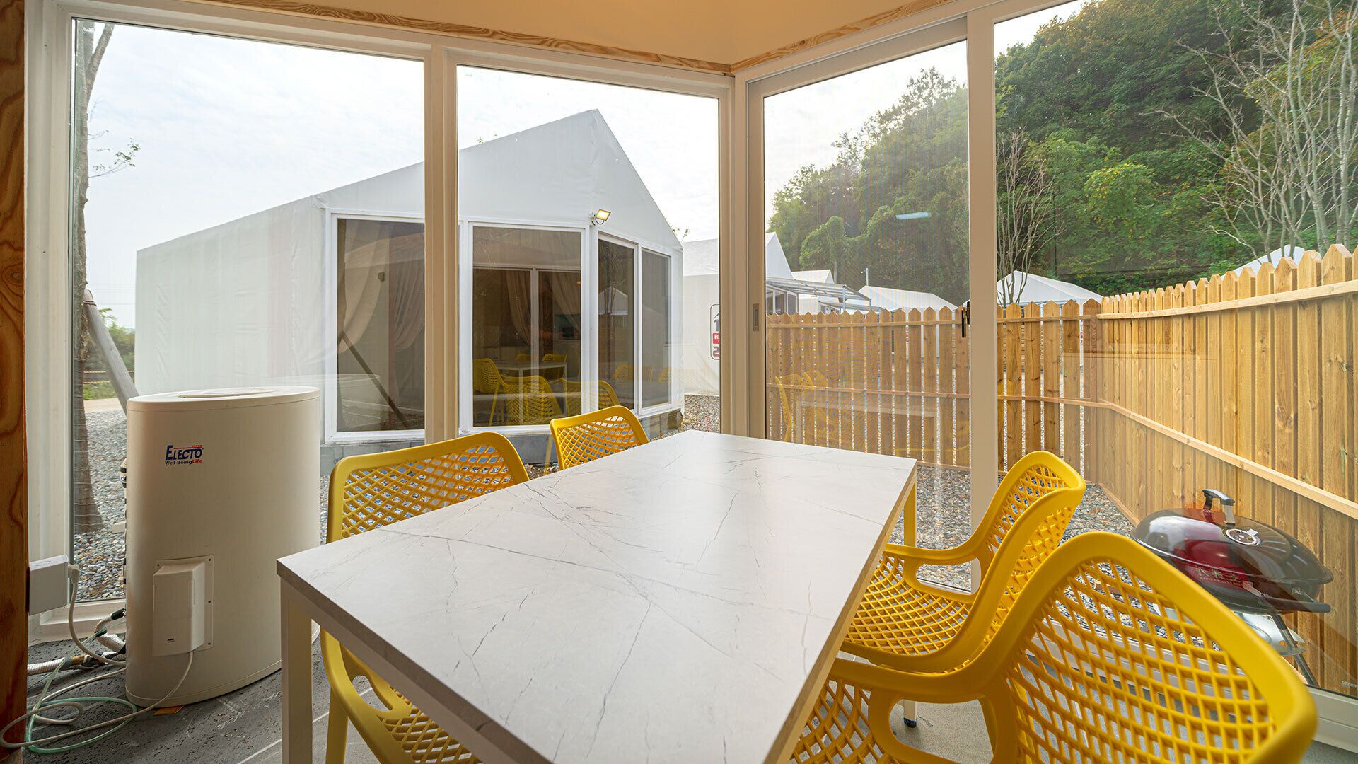 Een oranjerie met een witte tafel en gele stoelen, een witte verwarming en een houten hek op de achtergrond.